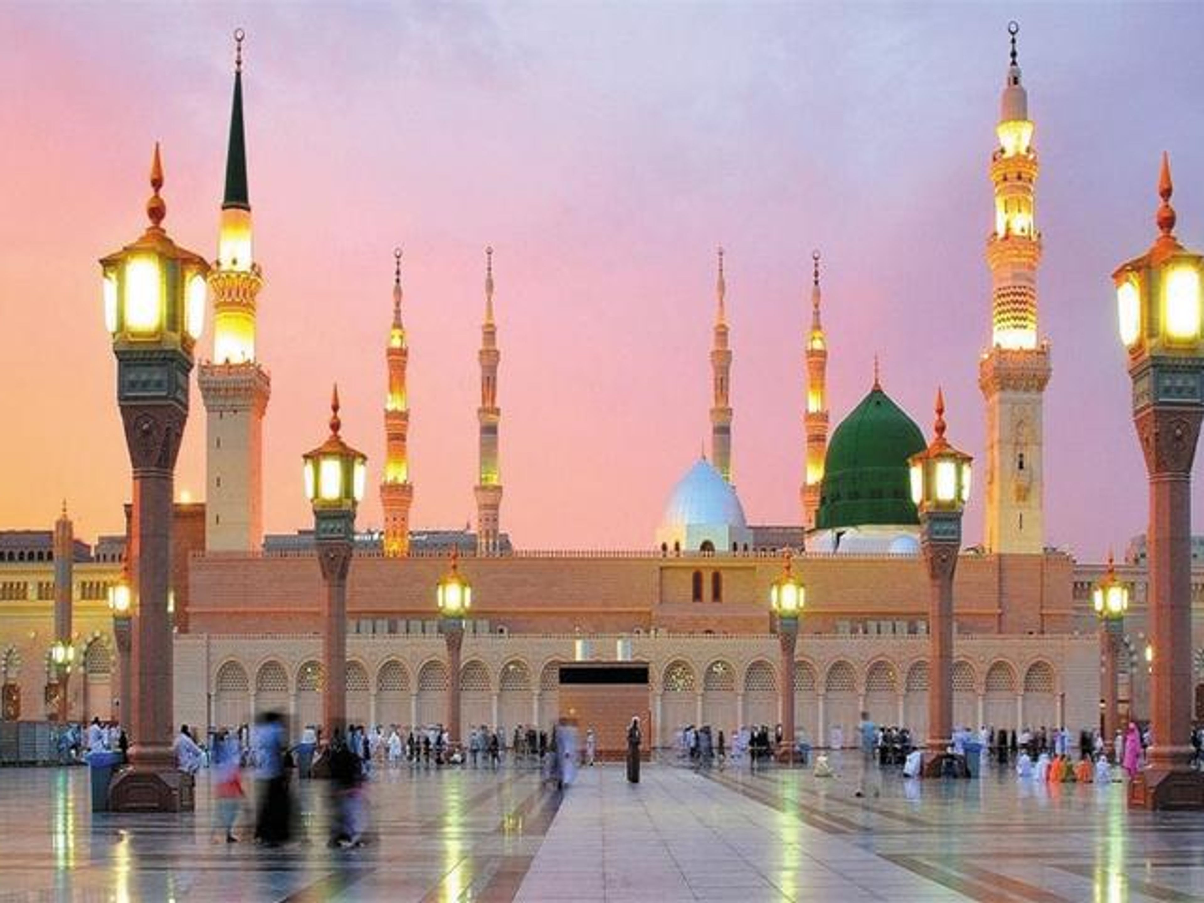 The Prophet's Mosque in Medina at twilight, with its green dome, illuminated minarets, and people in the courtyard.