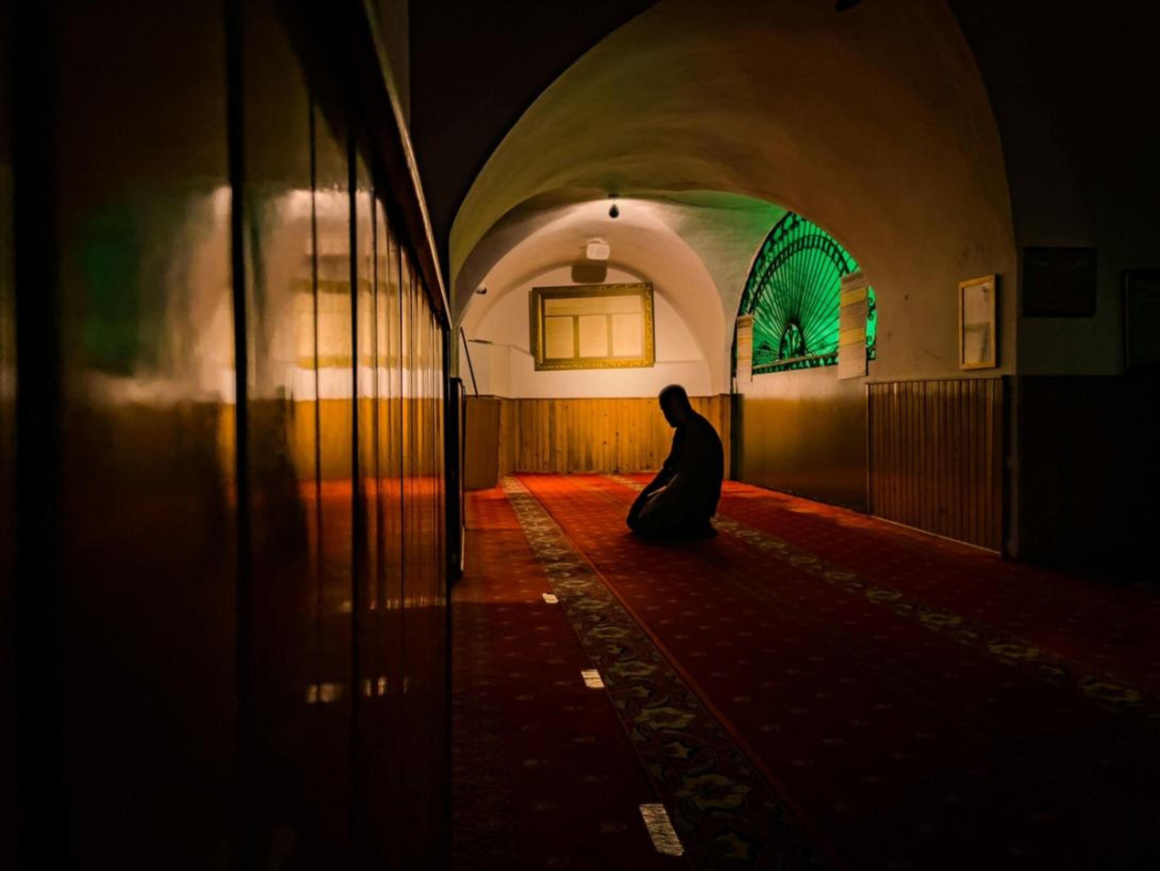 A person kneels in prayer on a red patterned carpet in a dimly lit, arched room with a bright green window.