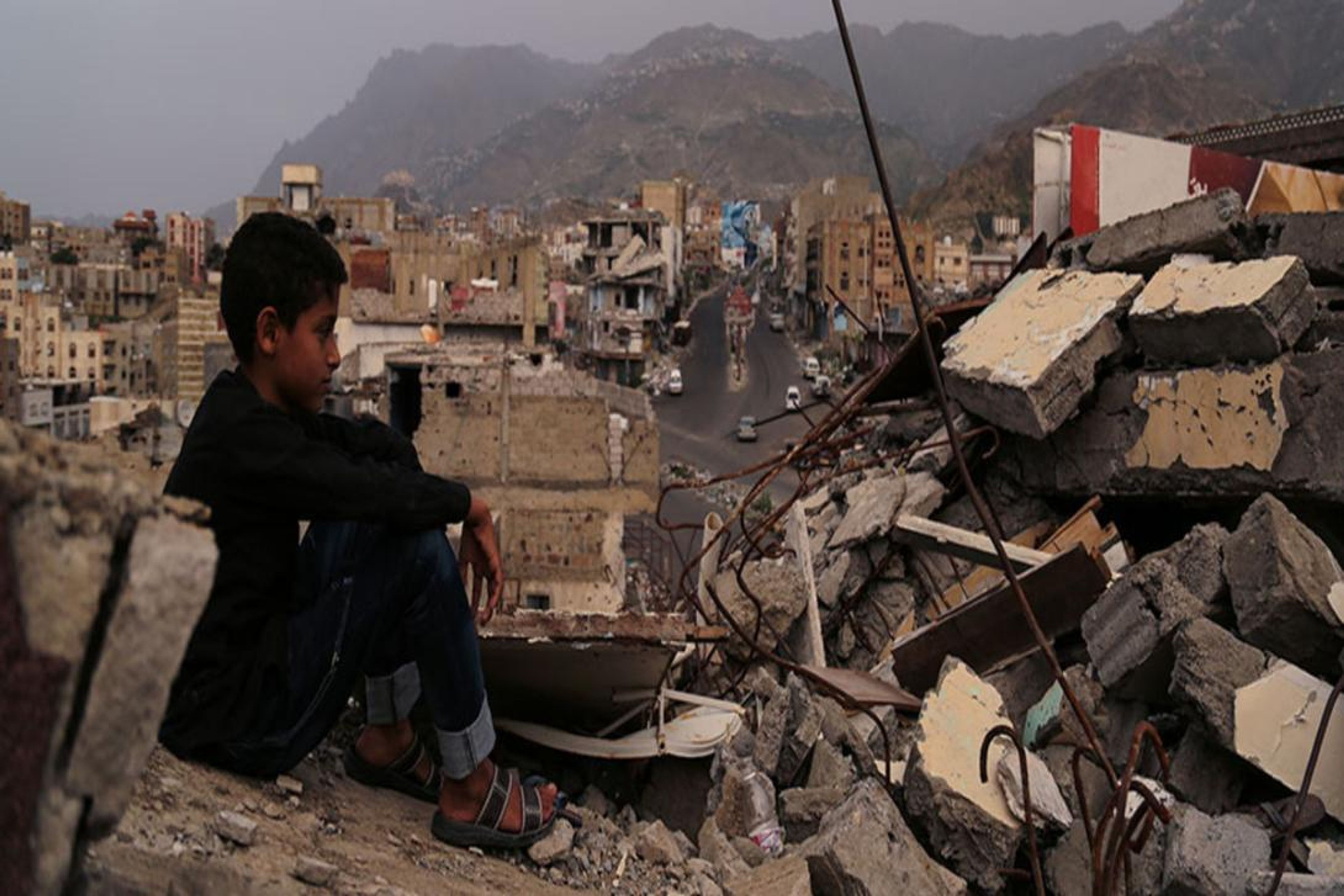 A pensive boy sits on rubble, looking out over a city largely destroyed by conflict.