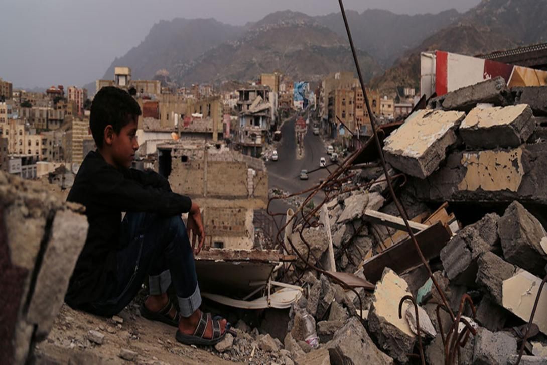 A pensive boy sits on rubble, looking out over a city largely destroyed by conflict.