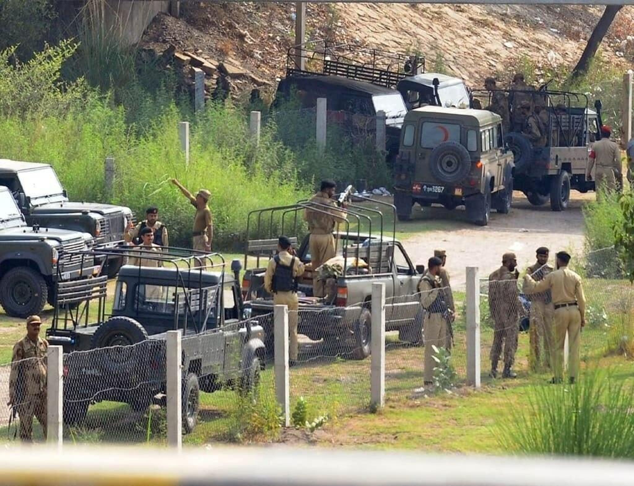 Uniformed personnel and multiple utility vehicles, one marked with a red cross, are gathered in a lush outdoor setting.