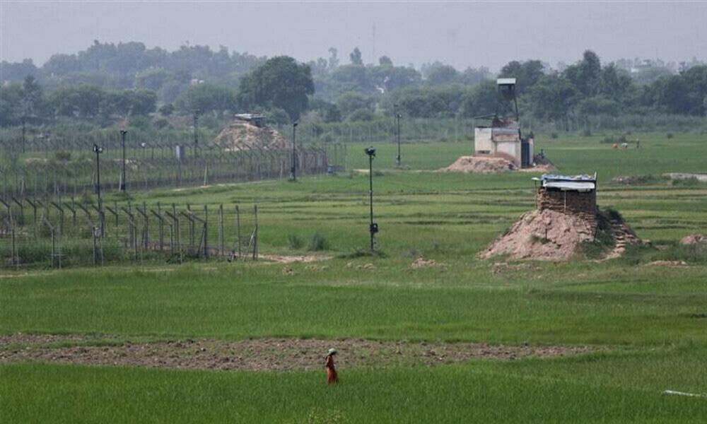 A border fence and watchtowers stretch across a green agricultural field with a person walking in the foreground.