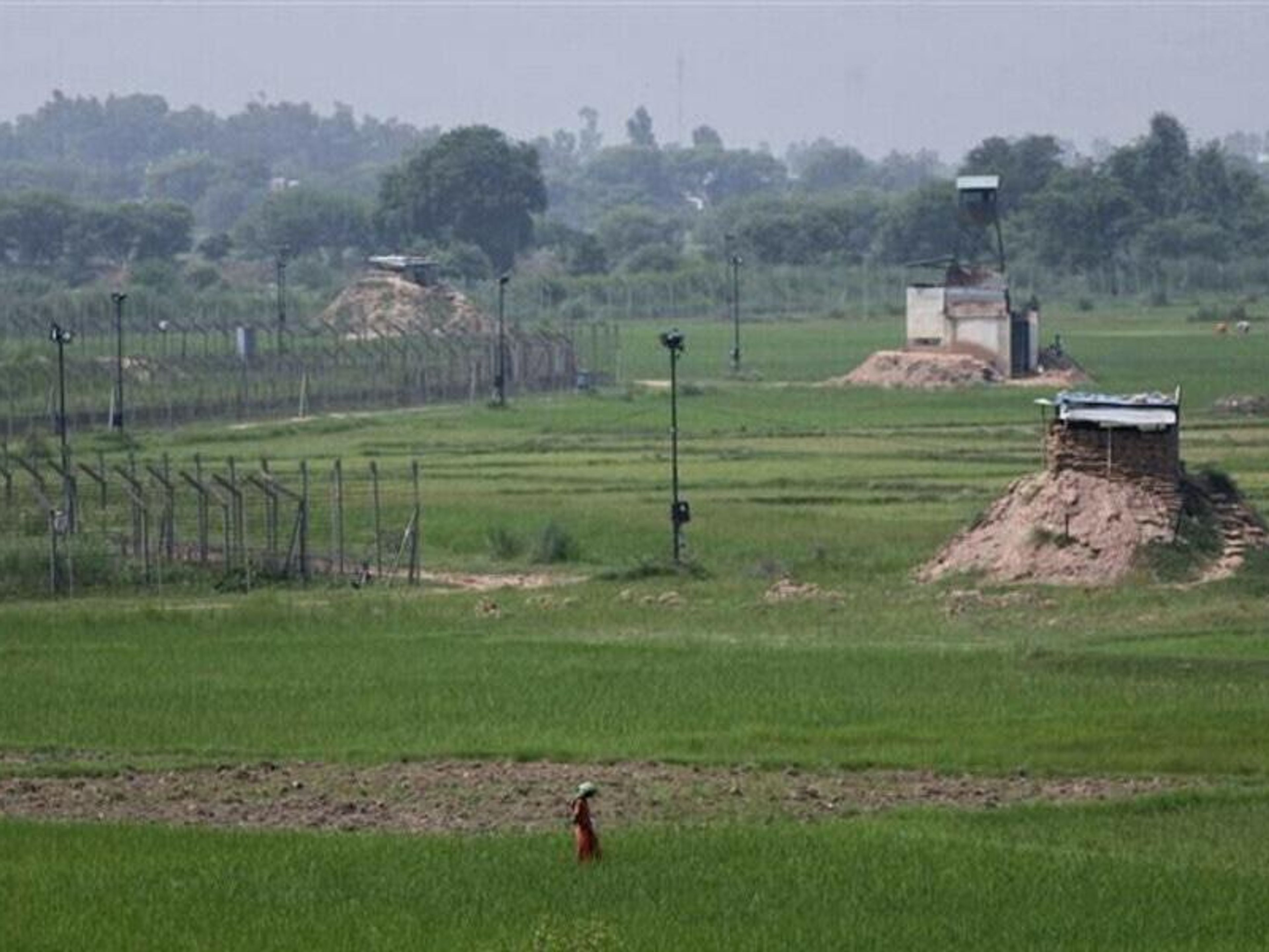 A border fence and watchtowers stretch across a green agricultural field with a person walking in the foreground.