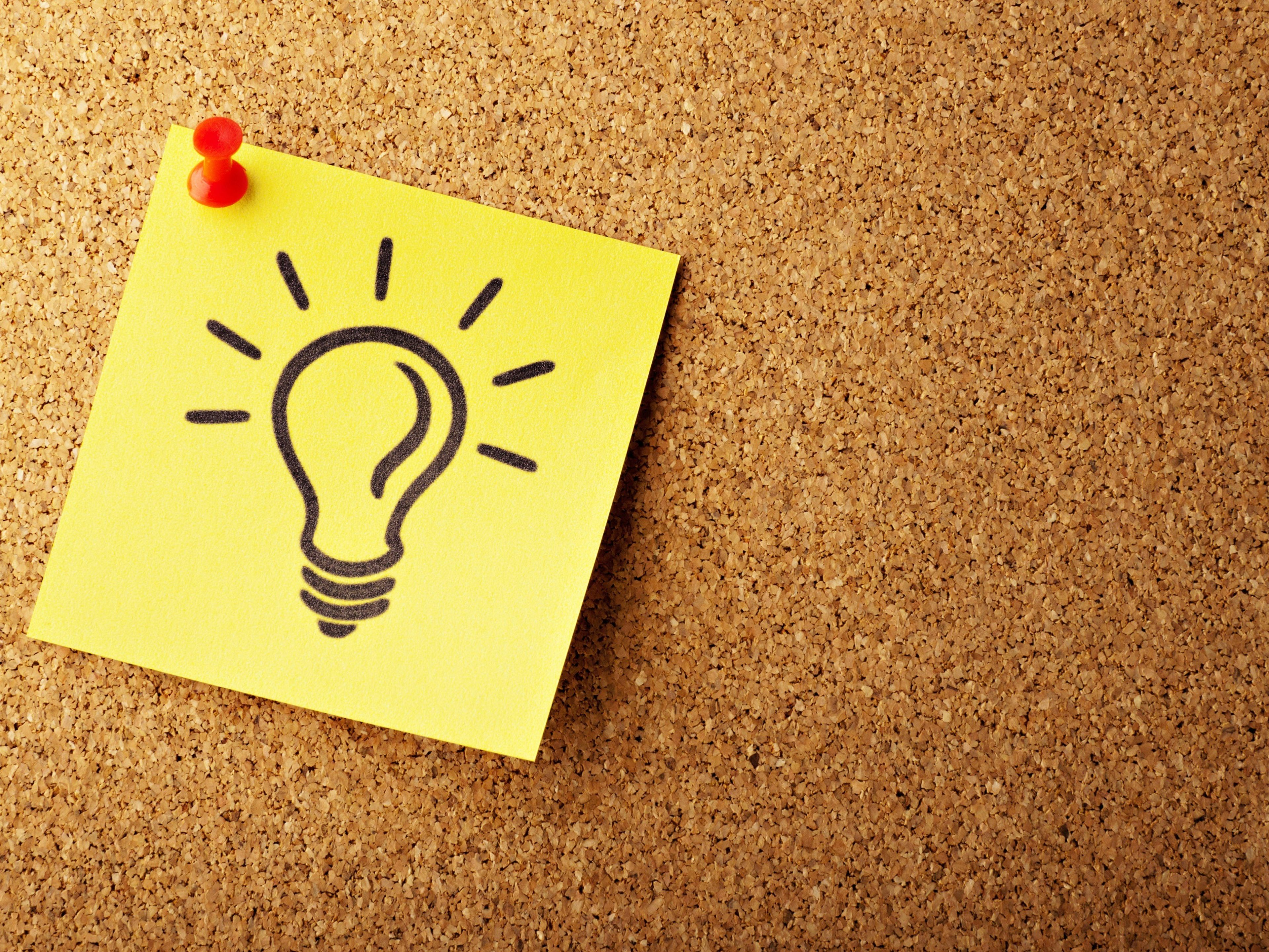 A yellow sticky note with a lightbulb drawing pinned to a cork board.