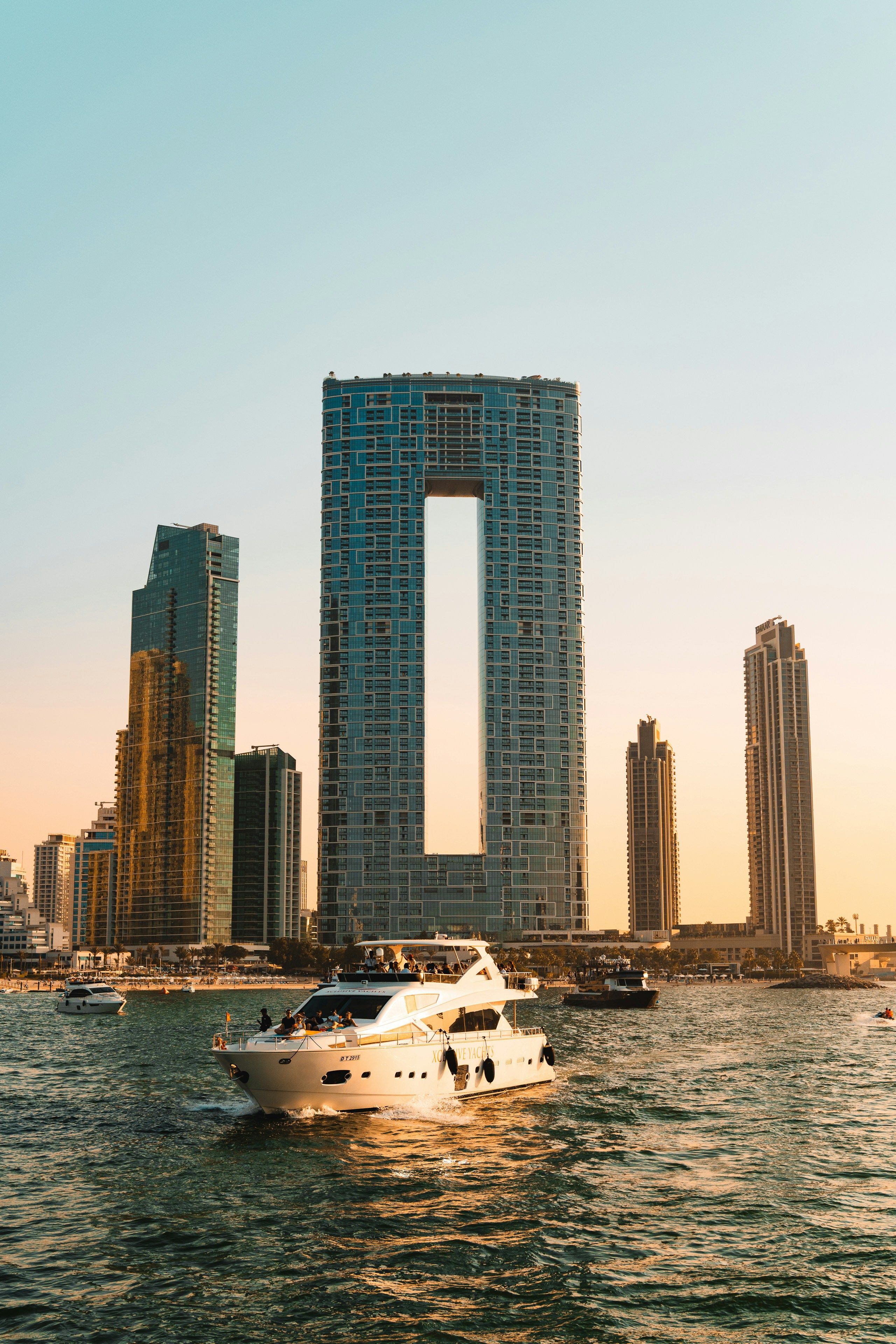A white yacht on the water with a city skyline, featuring a skyscraper with a large vertical cutout, at sunset.