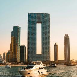 A white yacht on the water with a city skyline, featuring a skyscraper with a large vertical cutout, at sunset.