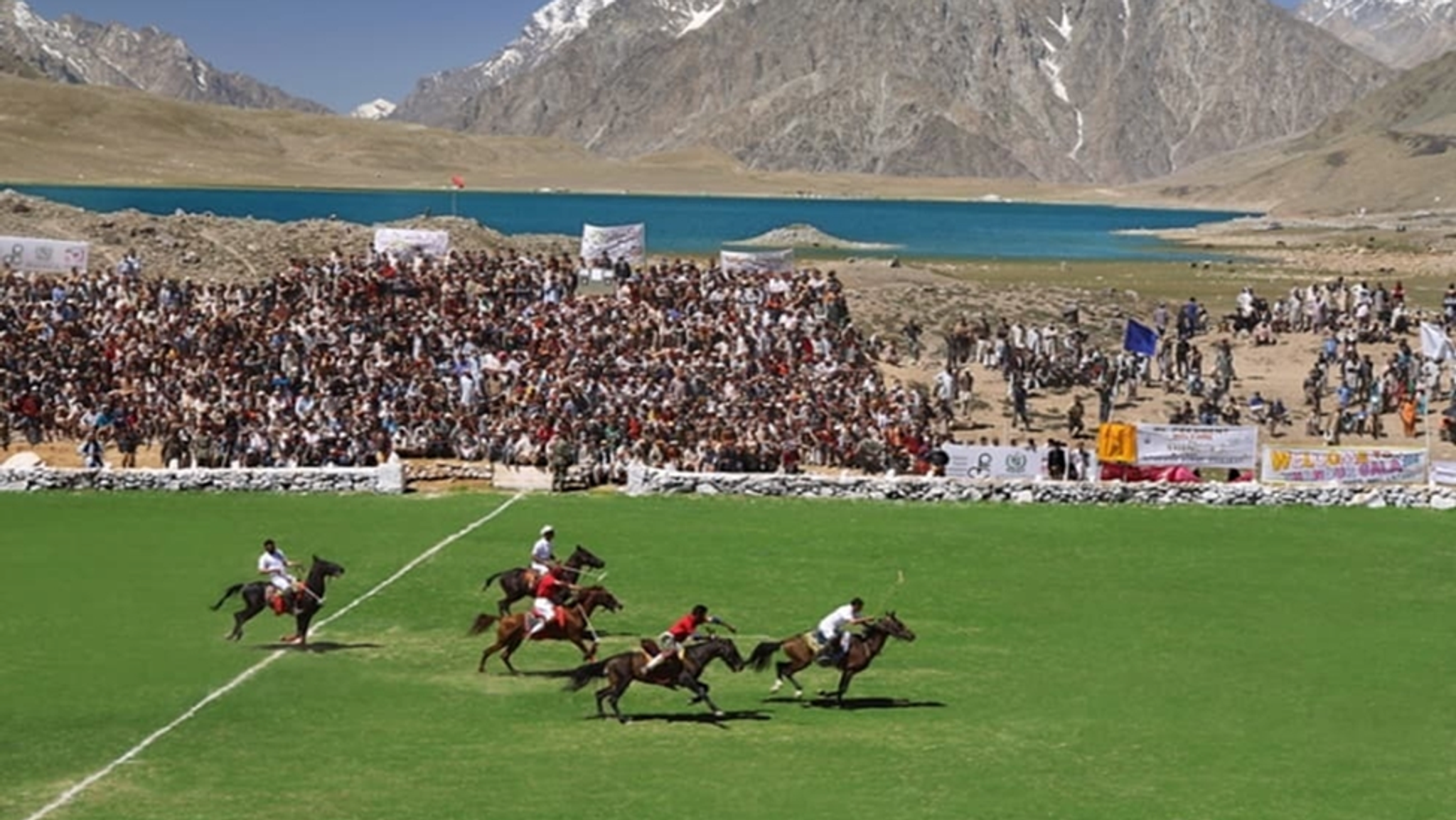 Polo match played on a green field before a large crowd, with a blue lake and snow-capped mountains in the background.