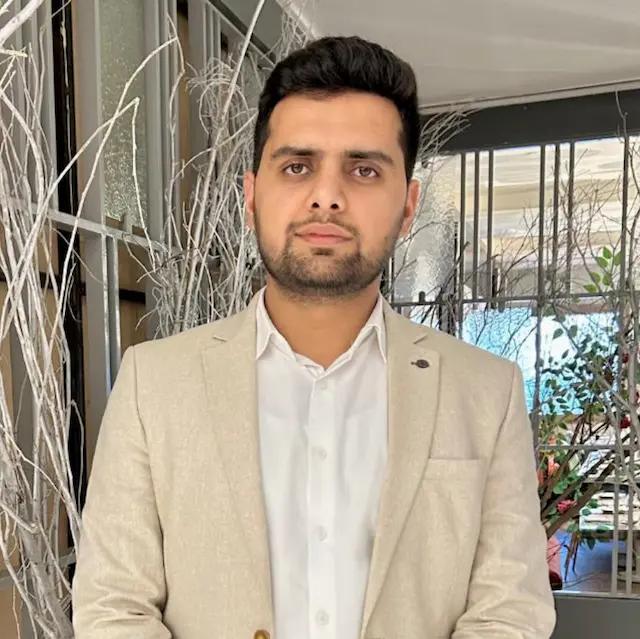 A man with dark hair and beard, wearing a beige blazer and white shirt.