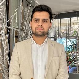 A man with dark hair and beard, wearing a beige blazer and white shirt.