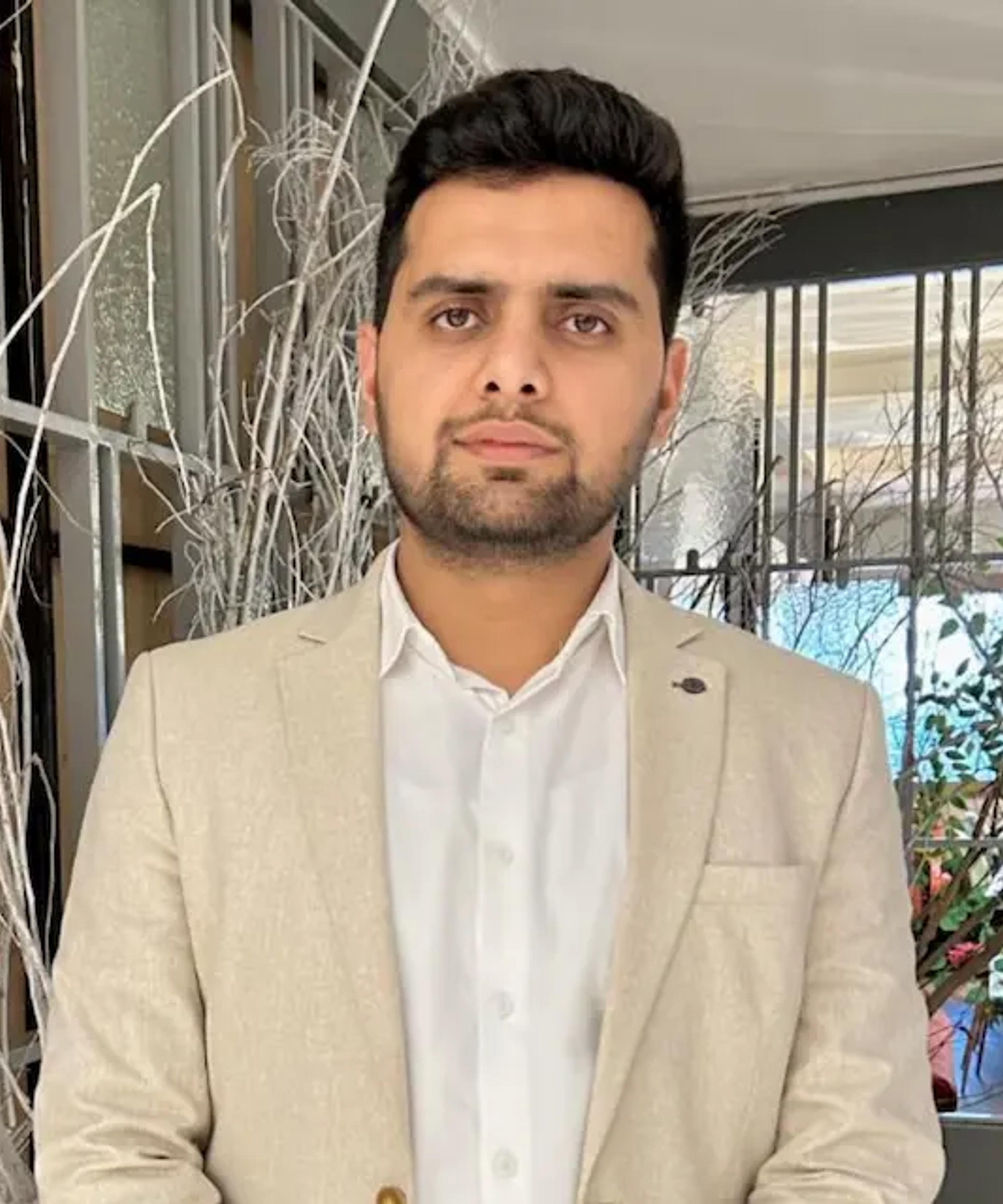 A man with dark hair and beard, wearing a beige blazer and white shirt.
