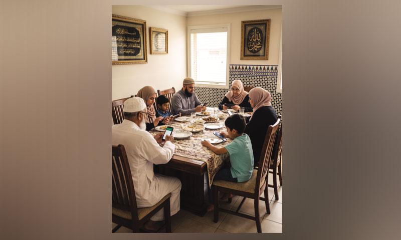 A multi-generational family sits at a dining table, with most members looking at their smartphones.