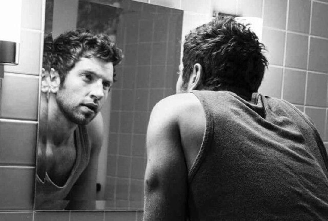 Black and white photo of a man from behind looking at his reflection in a mirror.