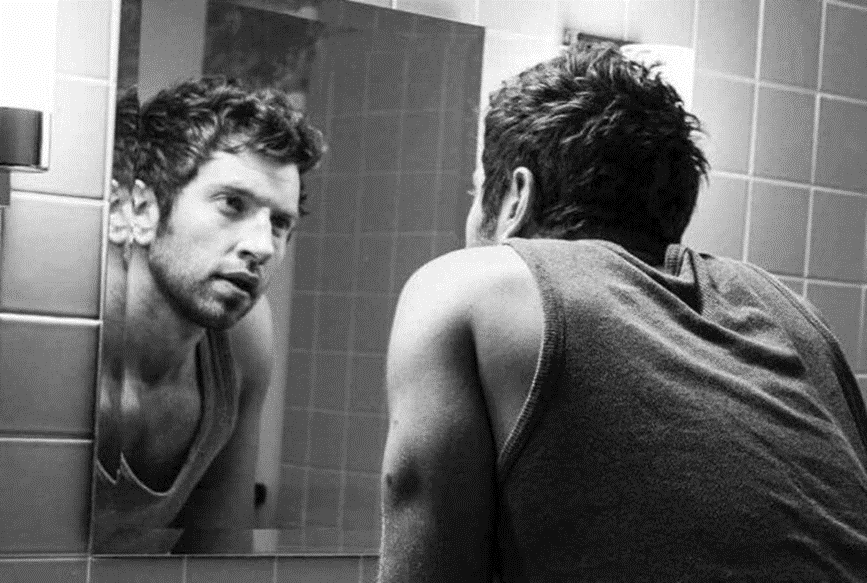 Black and white image of a man looking at his reflection in a mirror.