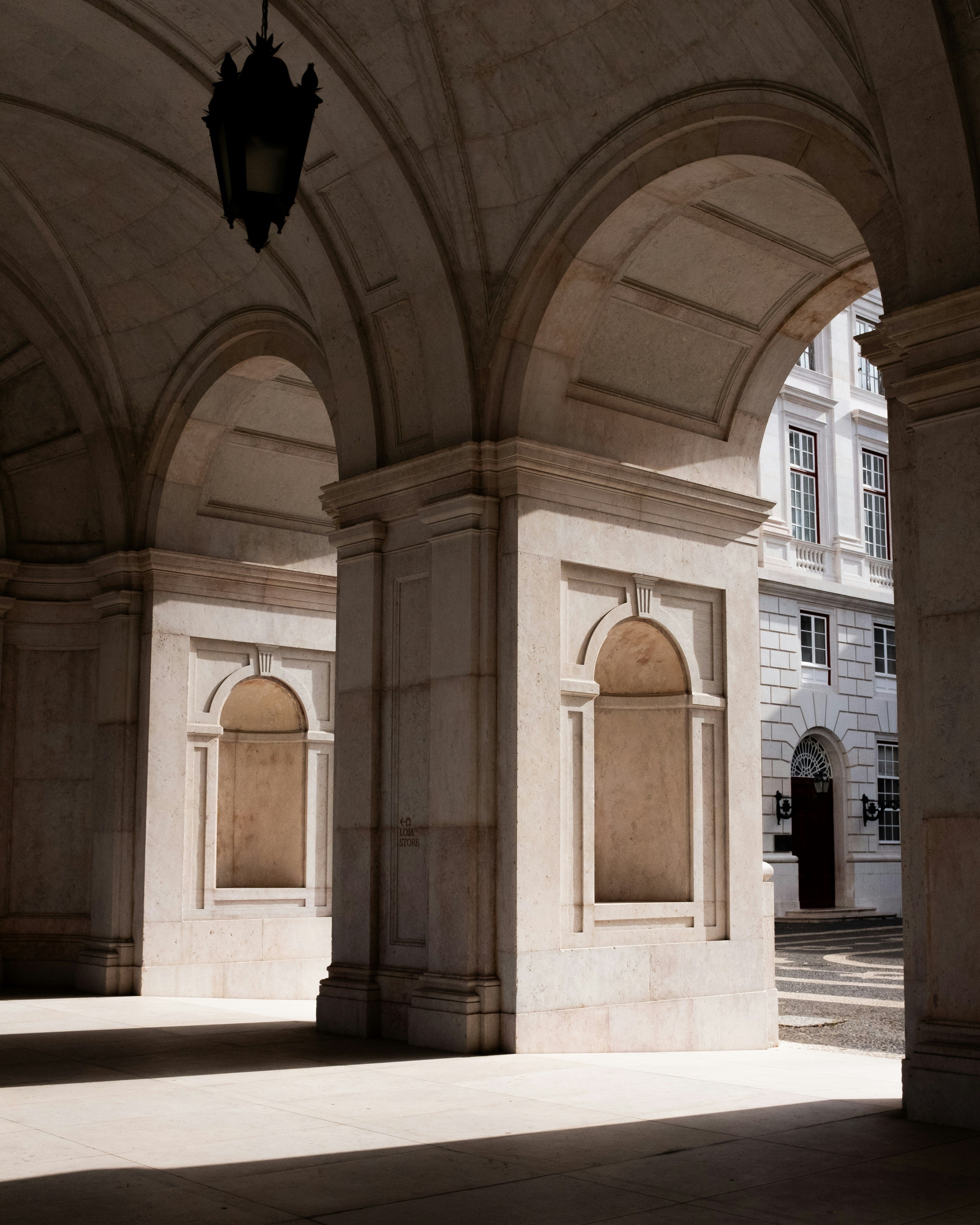 Stone arcade with a dark lantern, strong sunlight and shadows, and a white building in the background.