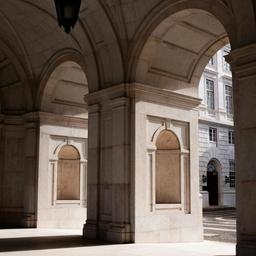 Stone arcade with a dark lantern, strong sunlight and shadows, and a white building in the background.