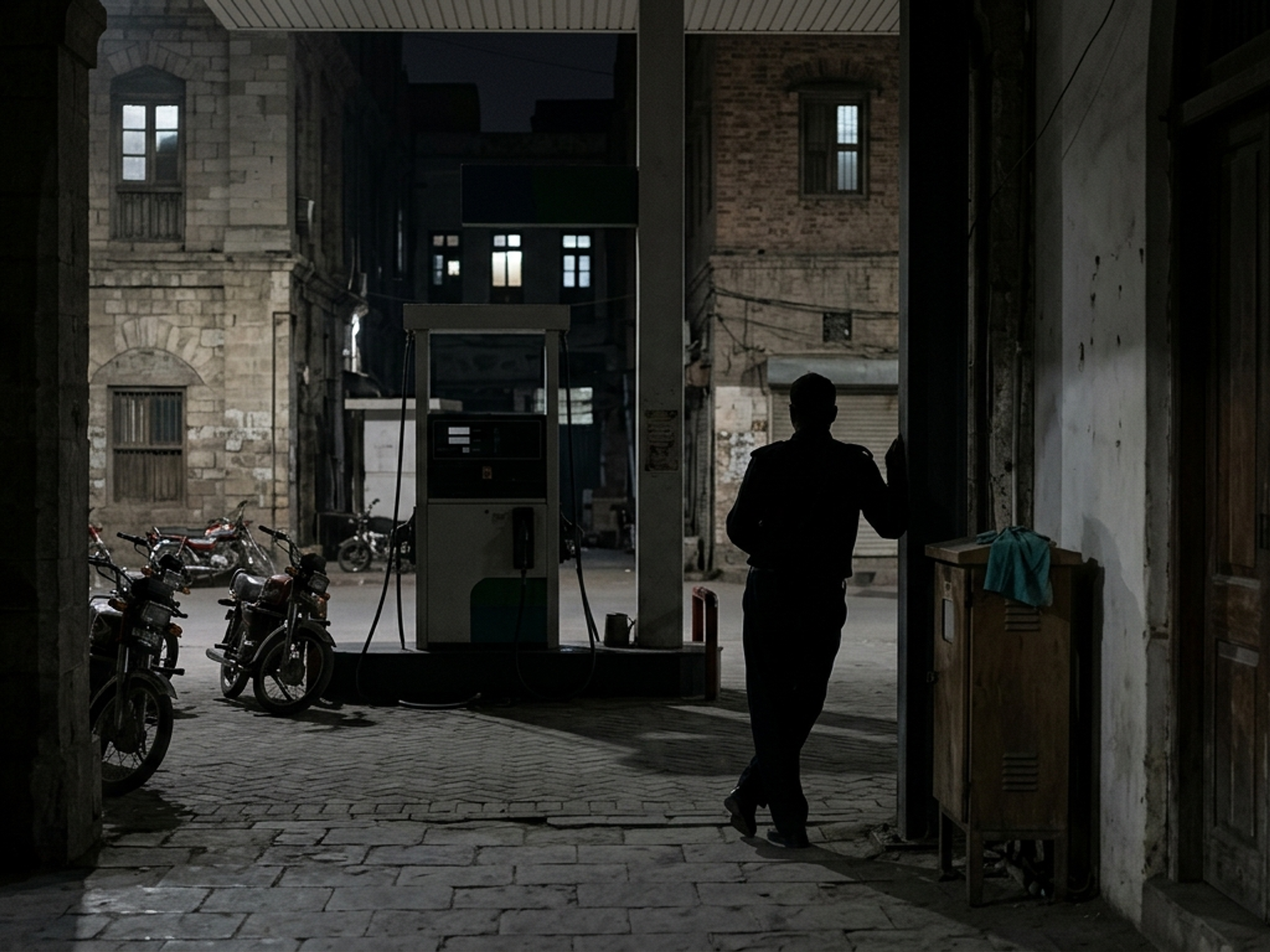 Silhouetted person leaning by a gas pump at night, with motorcycles and old buildings.