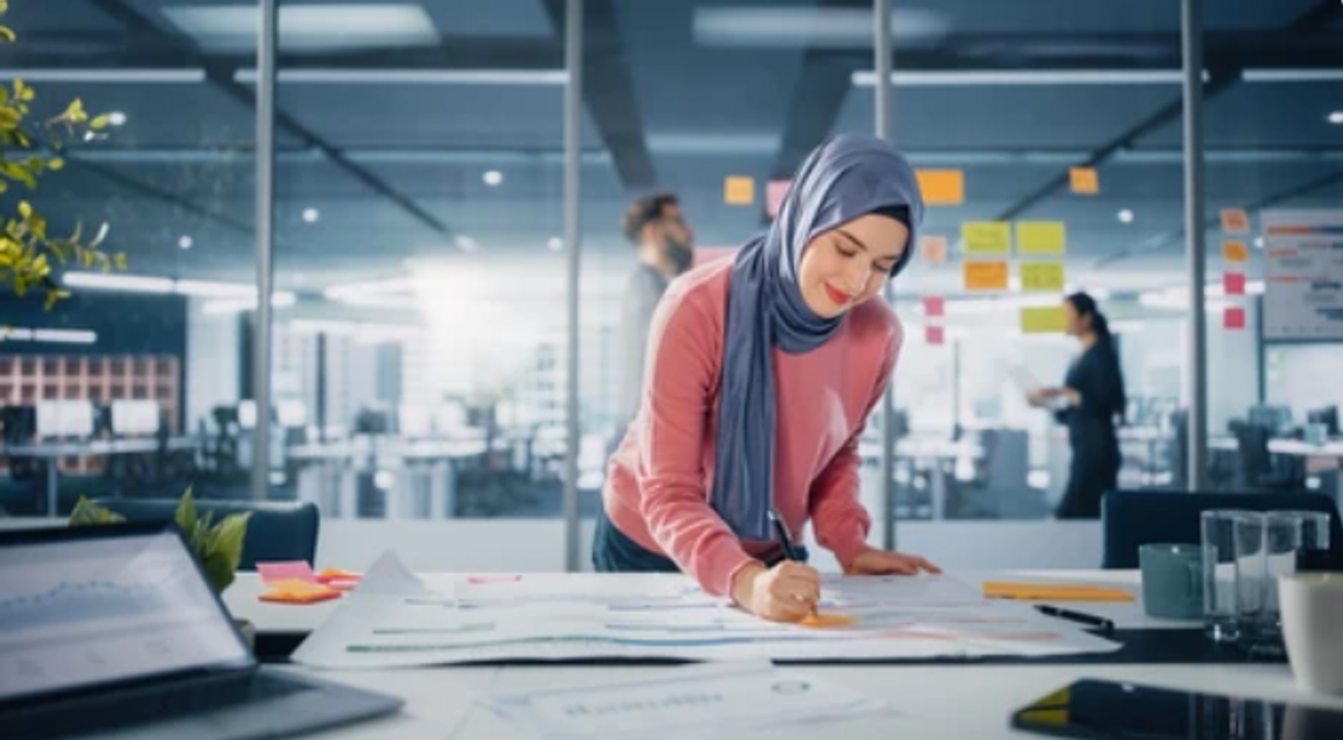 A smiling woman in a blue hijab writes on papers at a desk in a modern office.