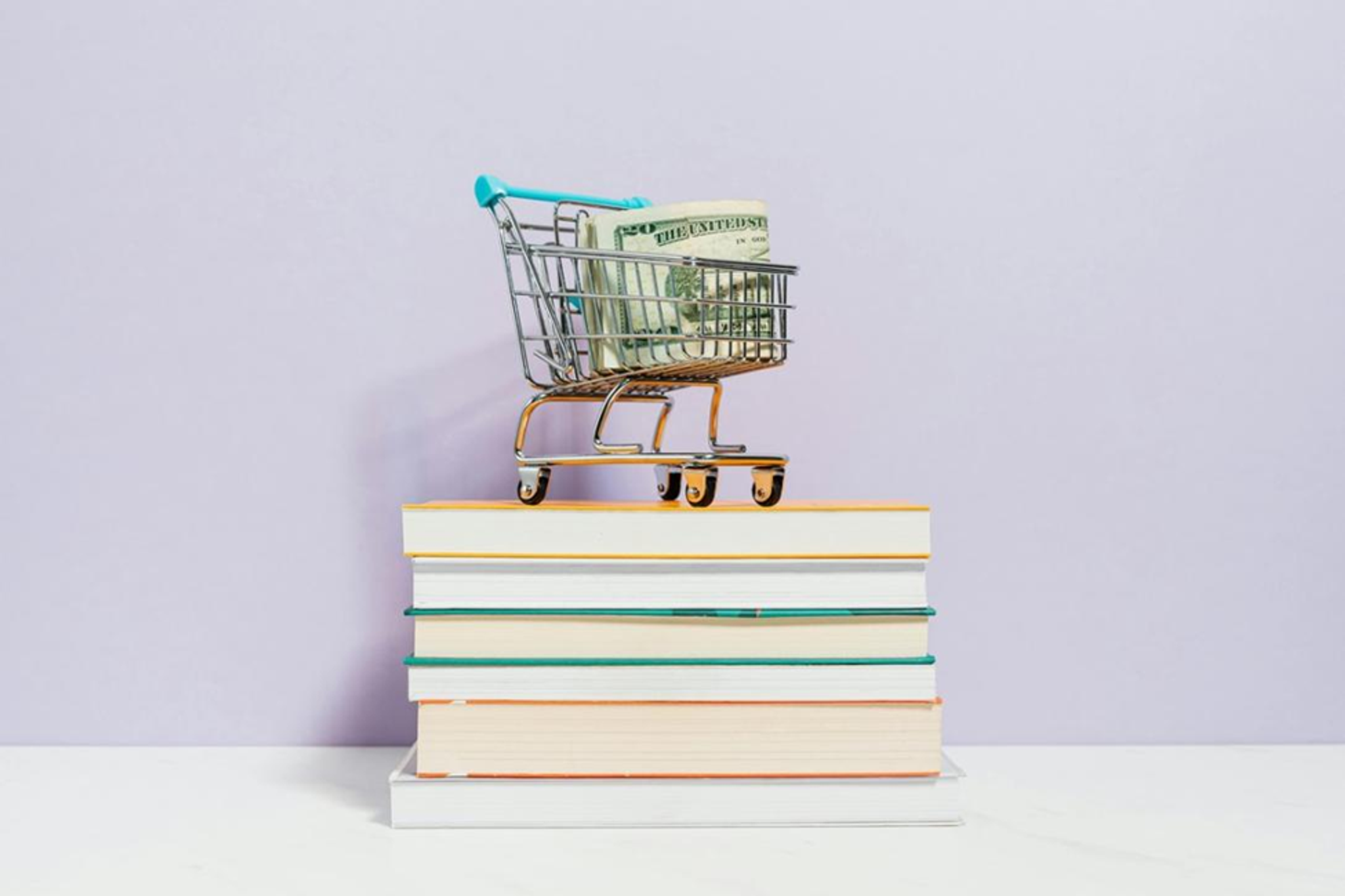 A miniature shopping cart with money sits on a stack of books.