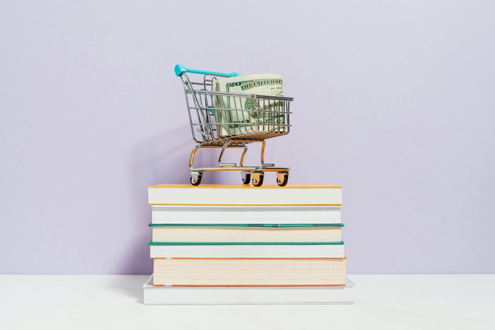 A miniature shopping cart with money sits on a stack of books.