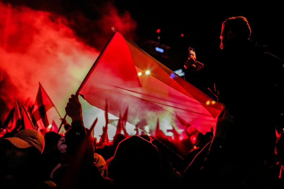 A nighttime crowd holding flags under red smoke and light, with a person filming.