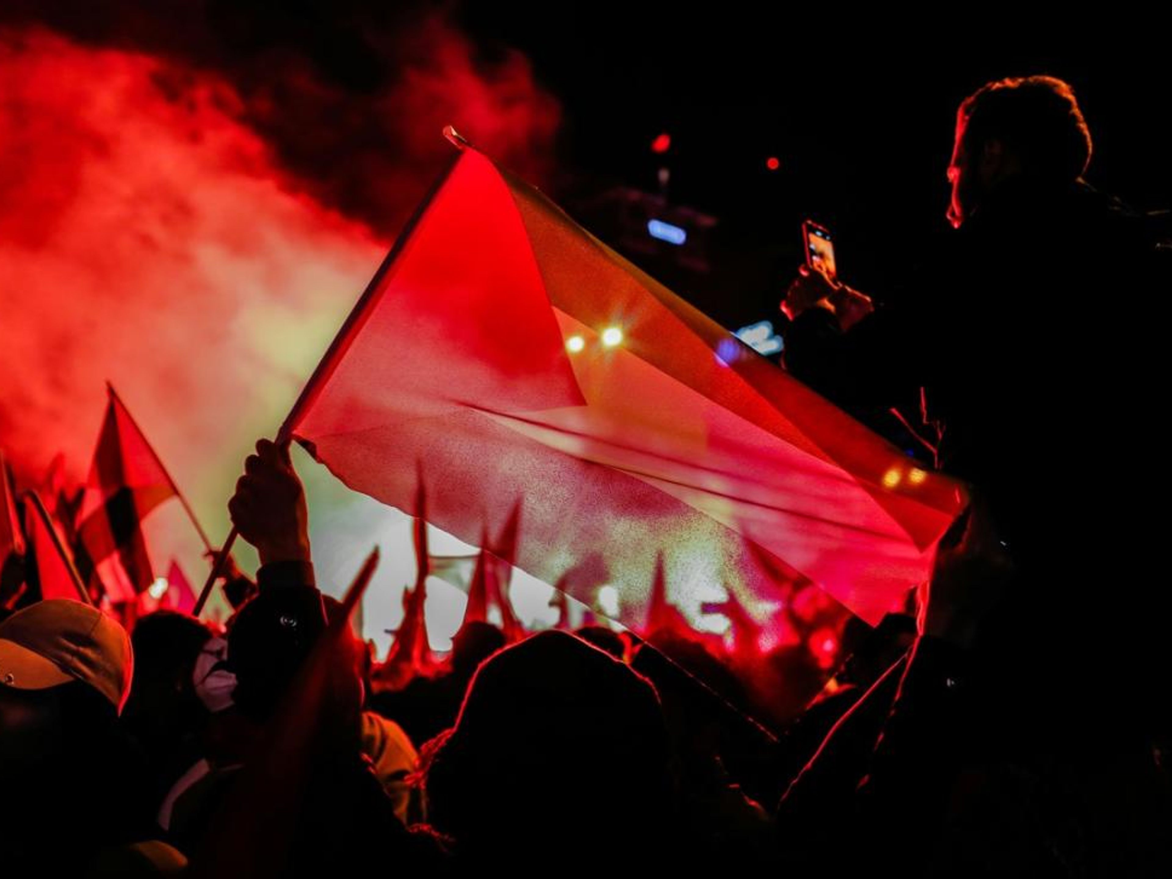 A nighttime crowd holding flags under red smoke and light, with a person filming.