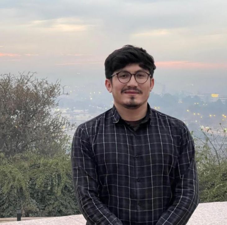 A young man in glasses and a dark plaid shirt smiles outdoors with a hazy city view in the background.