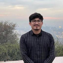 A young man in glasses and a dark plaid shirt smiles outdoors with a hazy city view in the background.