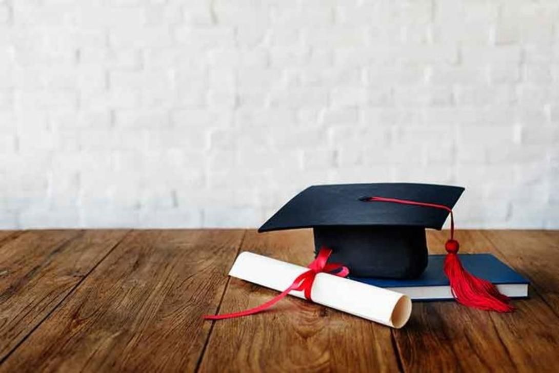 Black graduation cap with red tassel, rolled diploma with red ribbon, and a blue book on a wooden table.