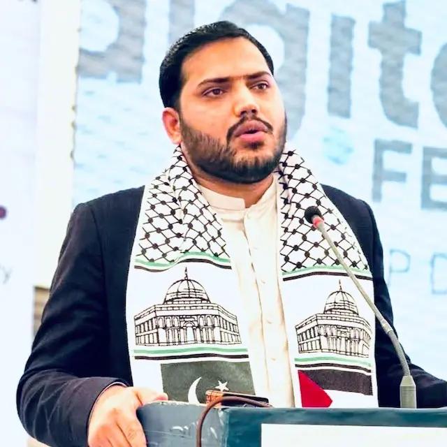 A man speaking into a microphone at a podium, wearing a scarf featuring the Dome of the Rock and the flags of Pakistan and Palestine.