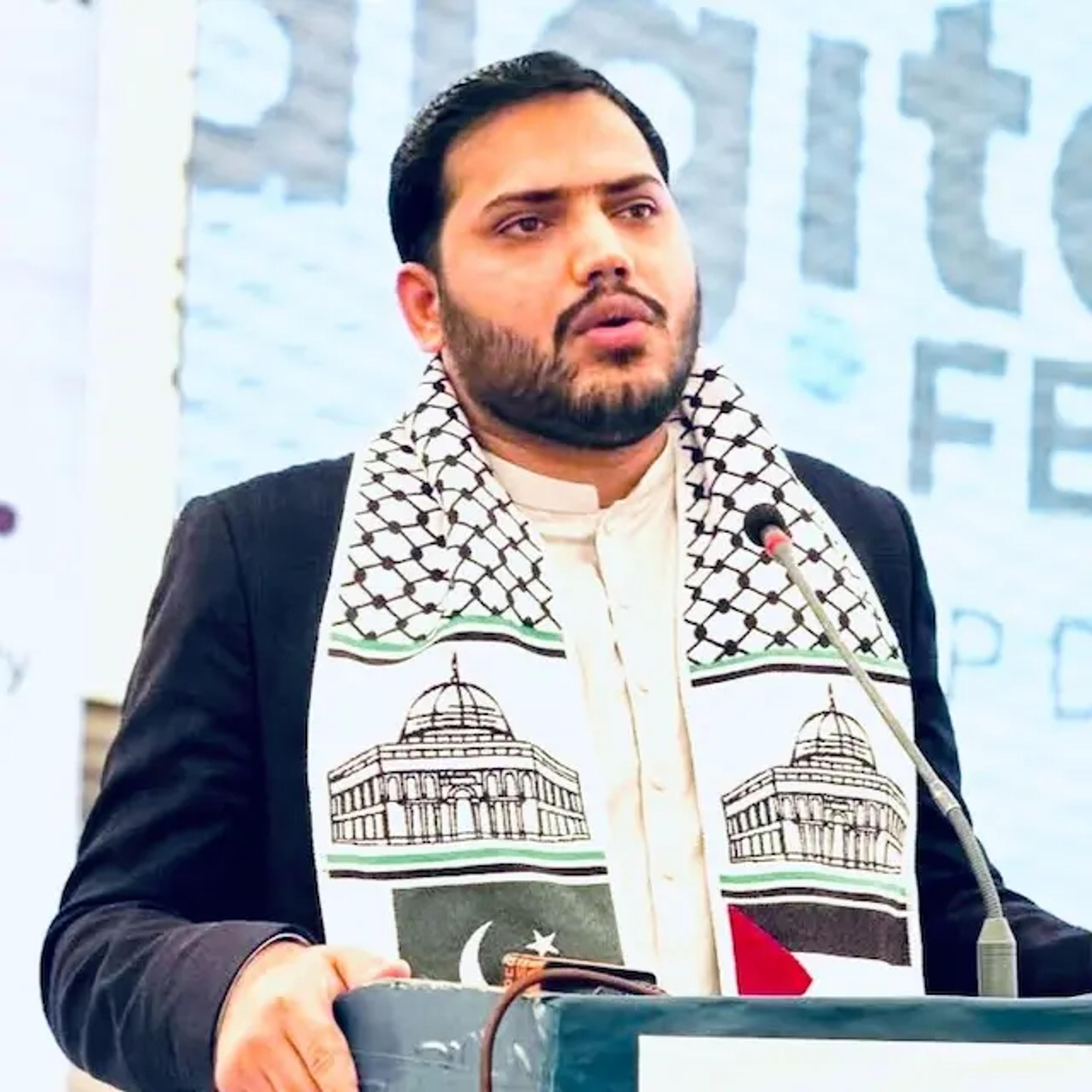 A man speaking into a microphone at a podium, wearing a scarf featuring the Dome of the Rock and the flags of Pakistan and Palestine.