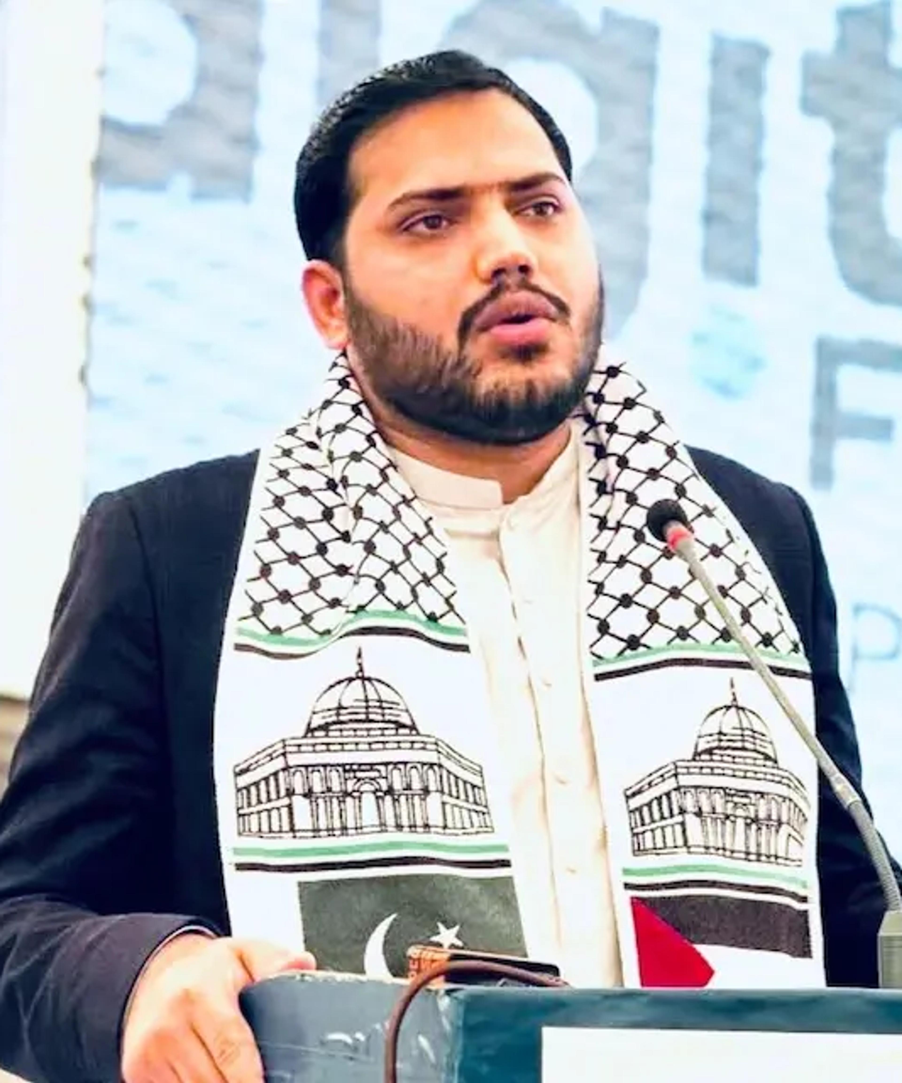 A man speaking into a microphone at a podium, wearing a scarf featuring the Dome of the Rock and the flags of Pakistan and Palestine.