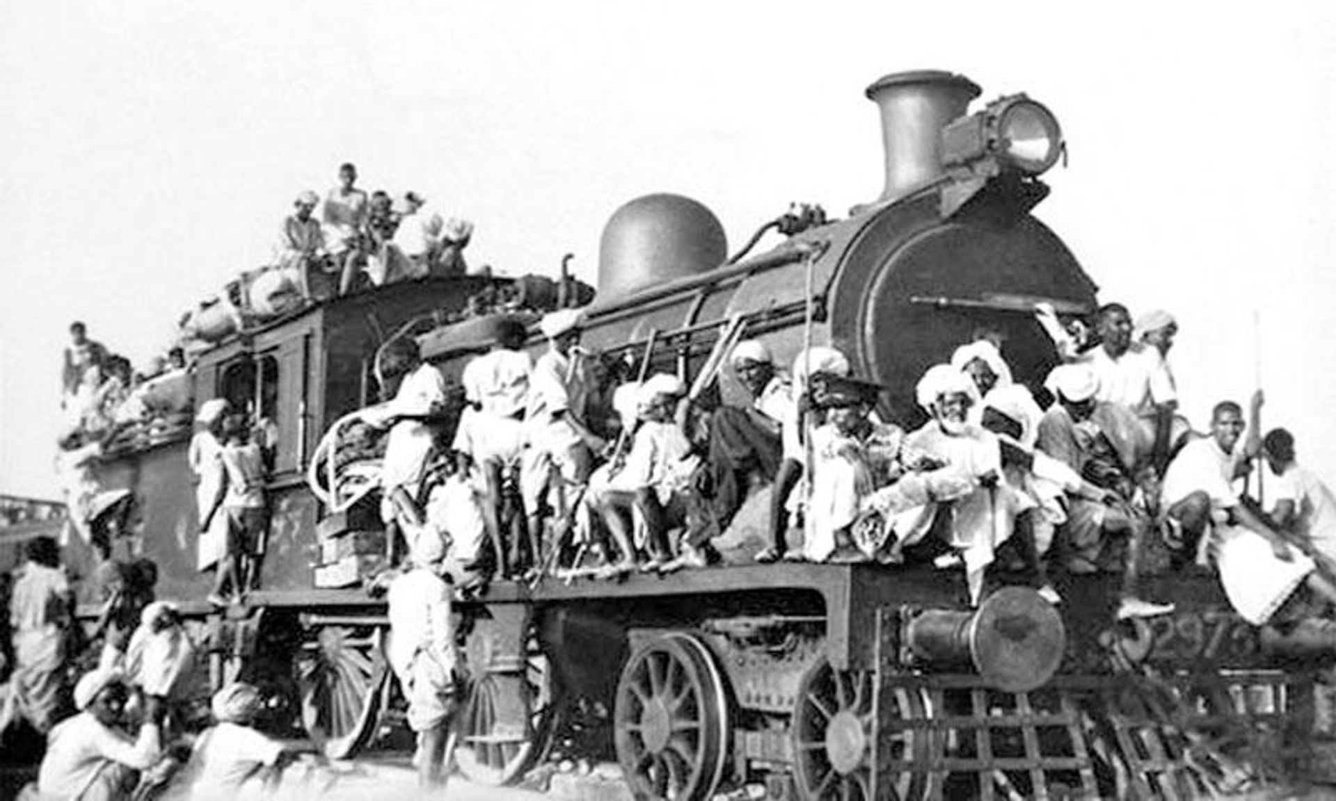 Black and white photo of a steam locomotive and tender heavily crowded with people riding on the roof, sides, and front.