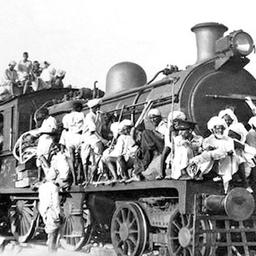Black and white photo of a steam train heavily crowded with people on its roof, sides, and front.
