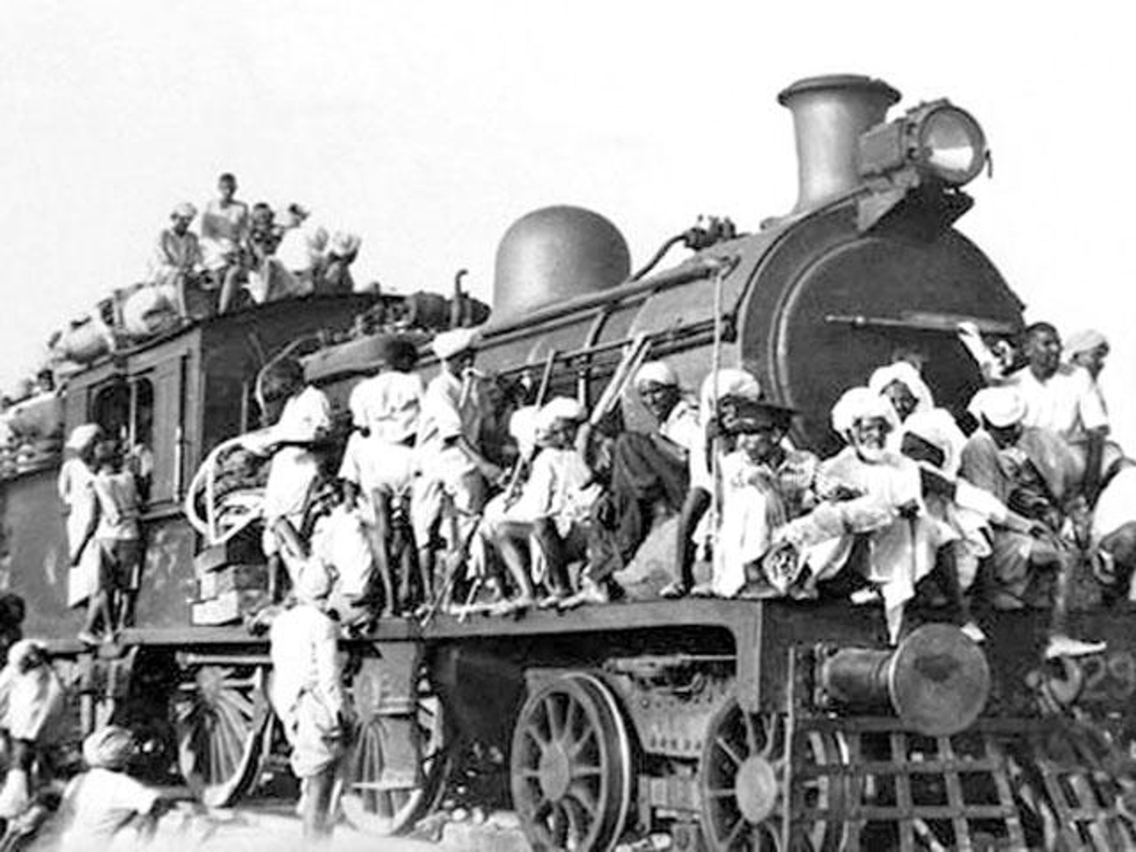 Black and white photo of a steam locomotive and tender heavily crowded with people riding on the roof, sides, and front.