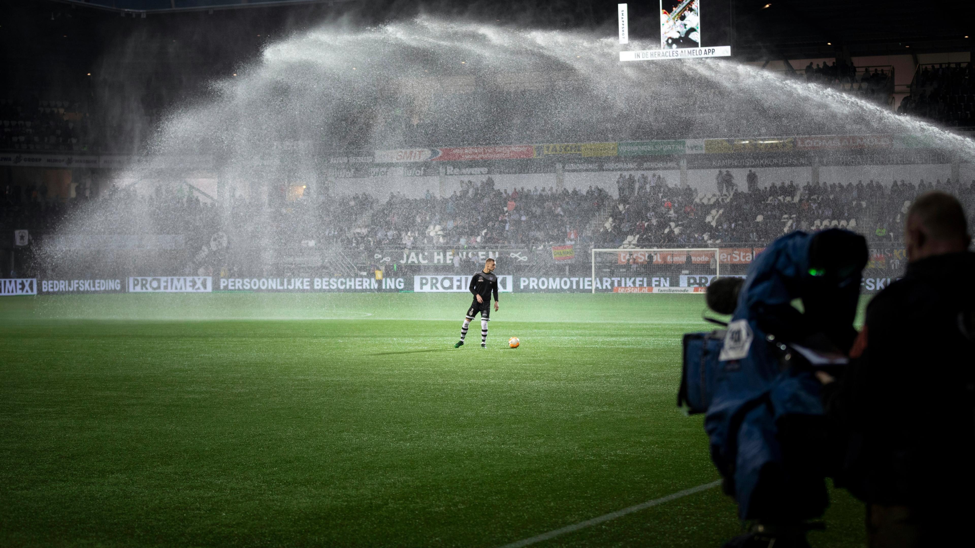 A soccer player on a field under a large arc of stadium sprinklers
