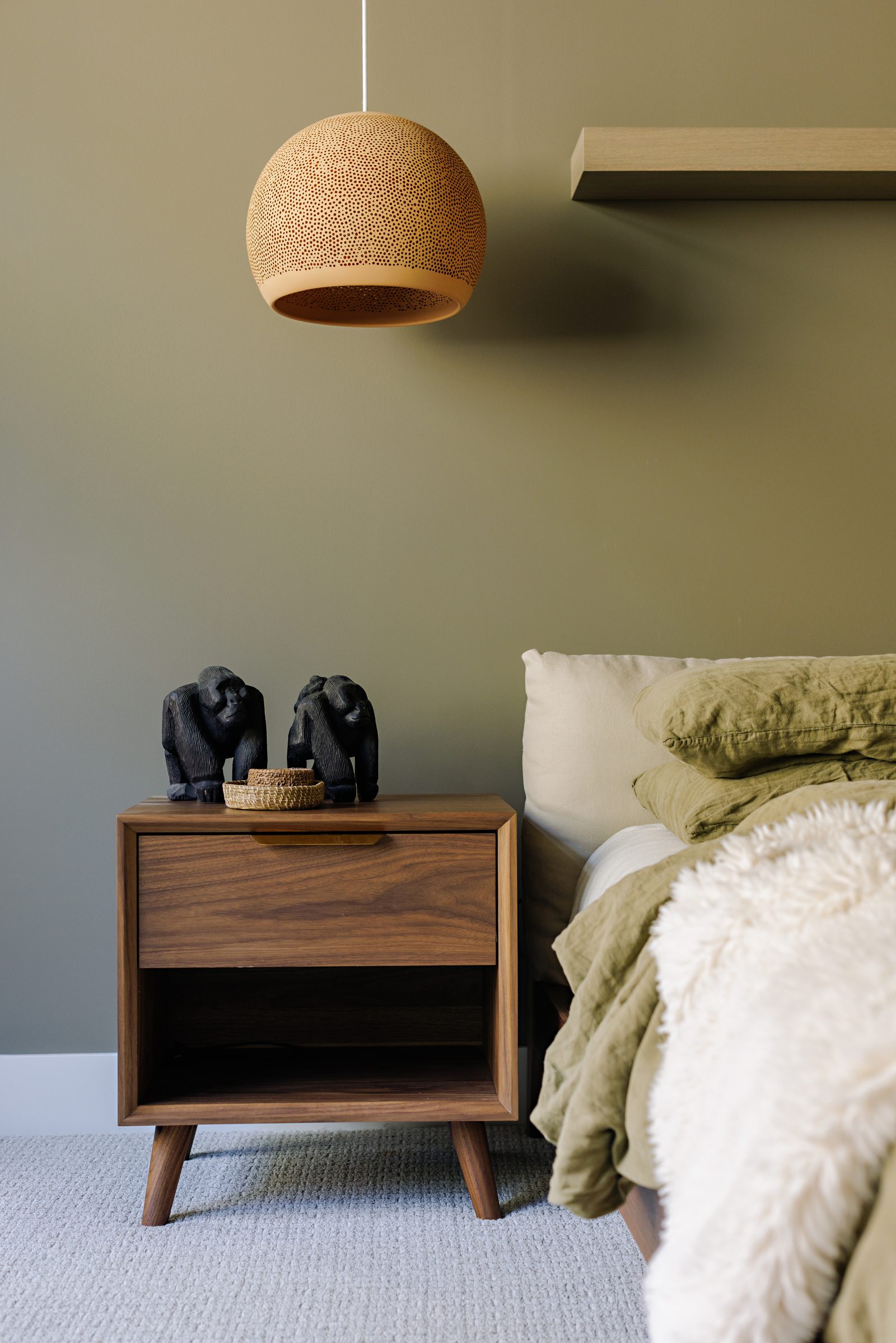 a walnut nightstand beneath a perforated terracotta pendant, two small carved figures and a woven dish on top.
