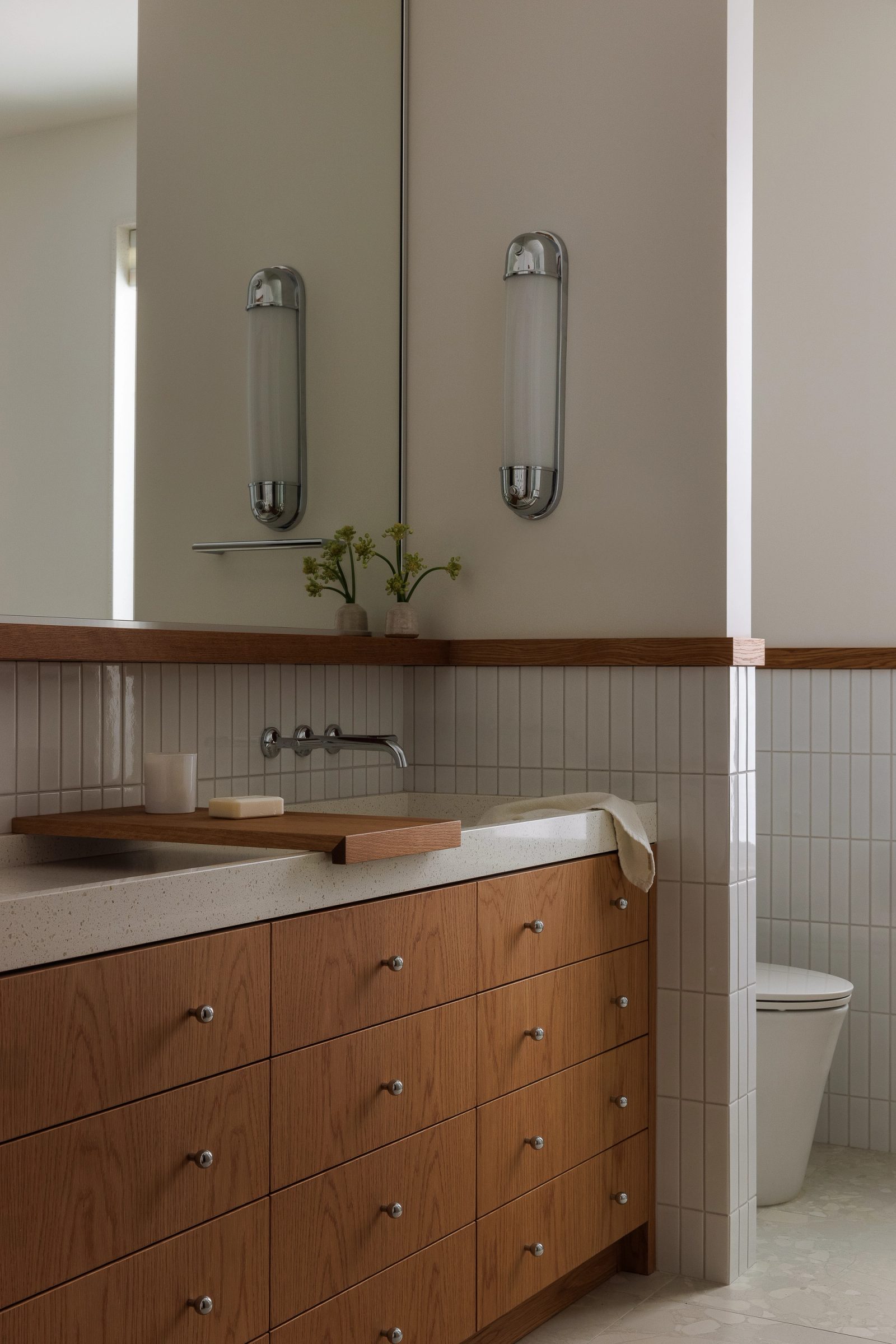 Left: a bathroom with tubular chrome sconces above a terrazzo counter, an oak shelf running the wall and fluted tile below.