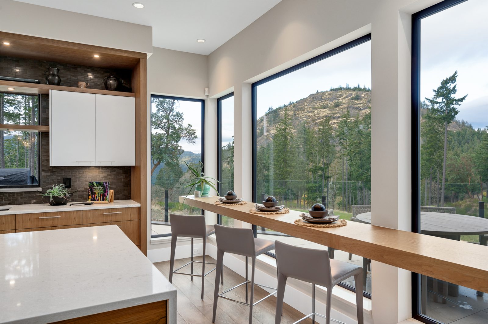 Oak counter and stools set against full-height glazing facing a bald rock bluff and firs