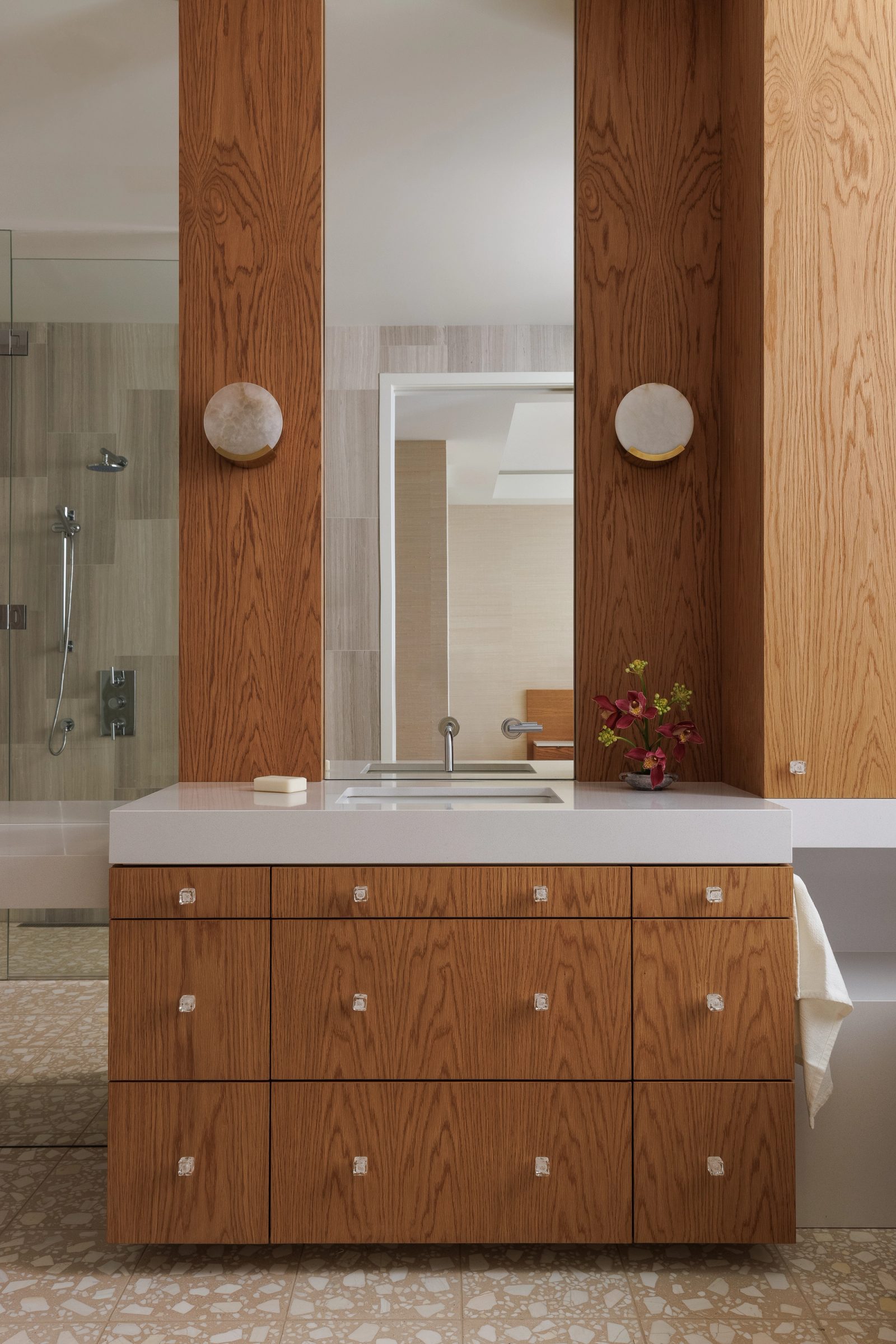 Left: a bathroom vanity in oak with glass cube pulls, alabaster discs to either side of a tall mirror, terrazzo underfoot.