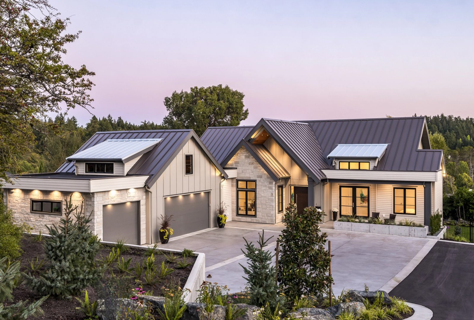 Estate home at dusk with a standing-seam metal roof, stone piers and lit windows above a concrete driveway