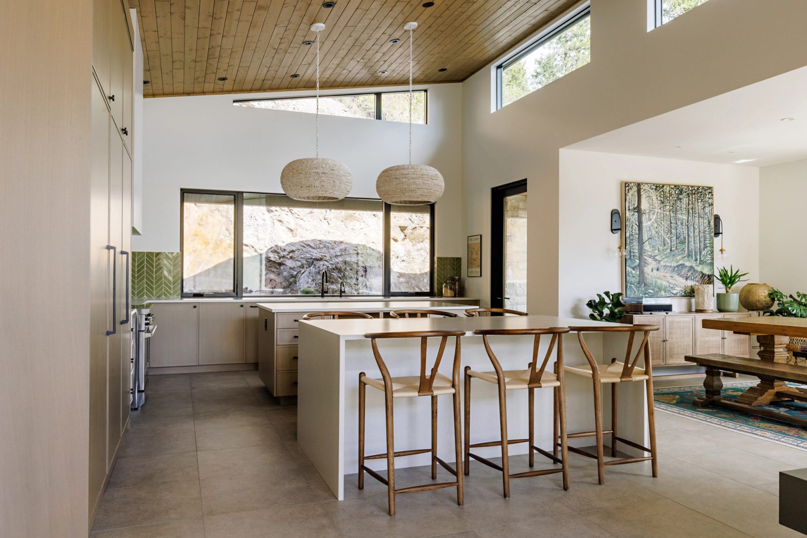 A kitchen with two pale wood islands under woven pendant shades, a band of clerestory glazing above, and a window over the sink filled entirely by rock.