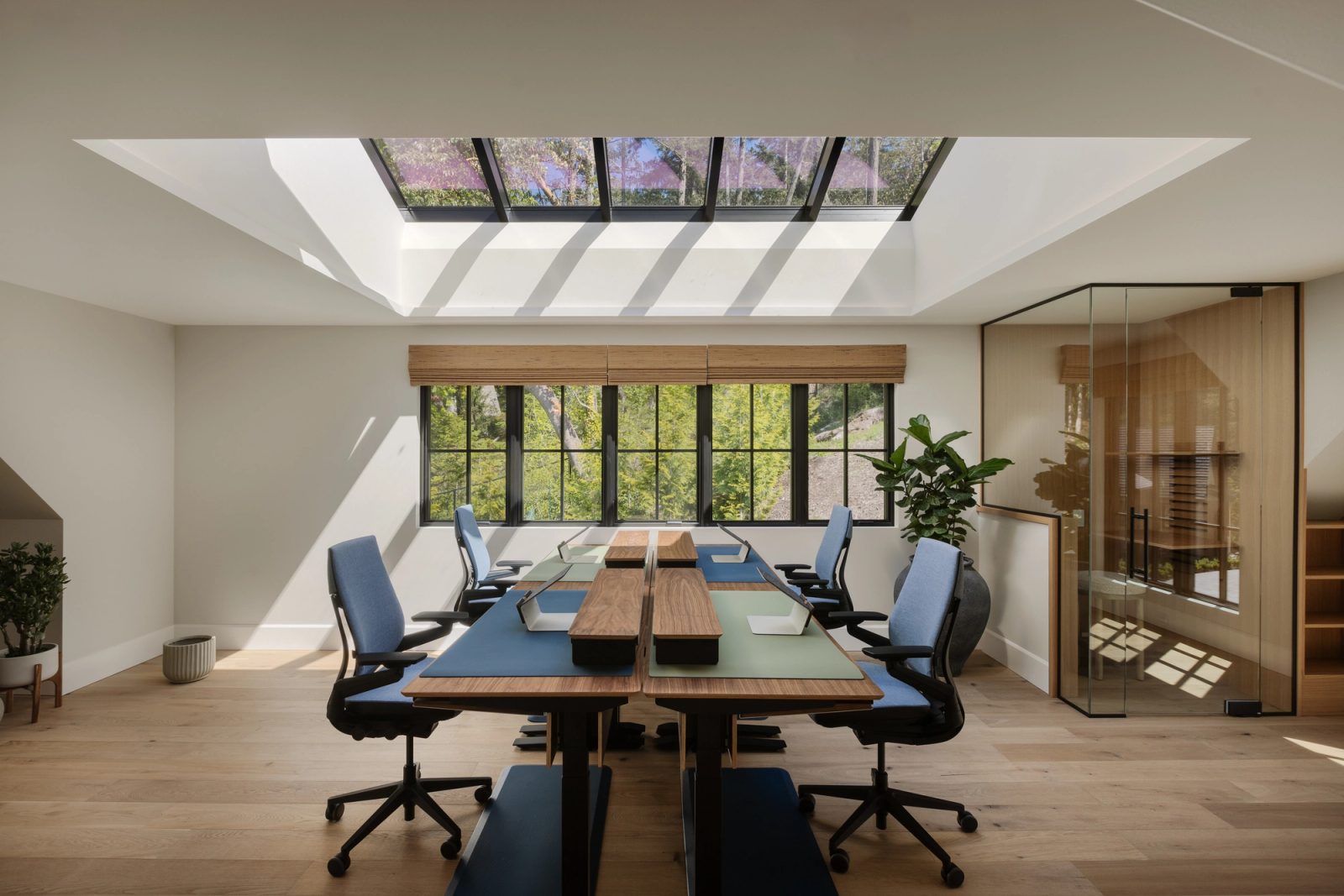 A study under a long ridge skylight, desks in a row facing windows to the forest and a glazed room beyond.