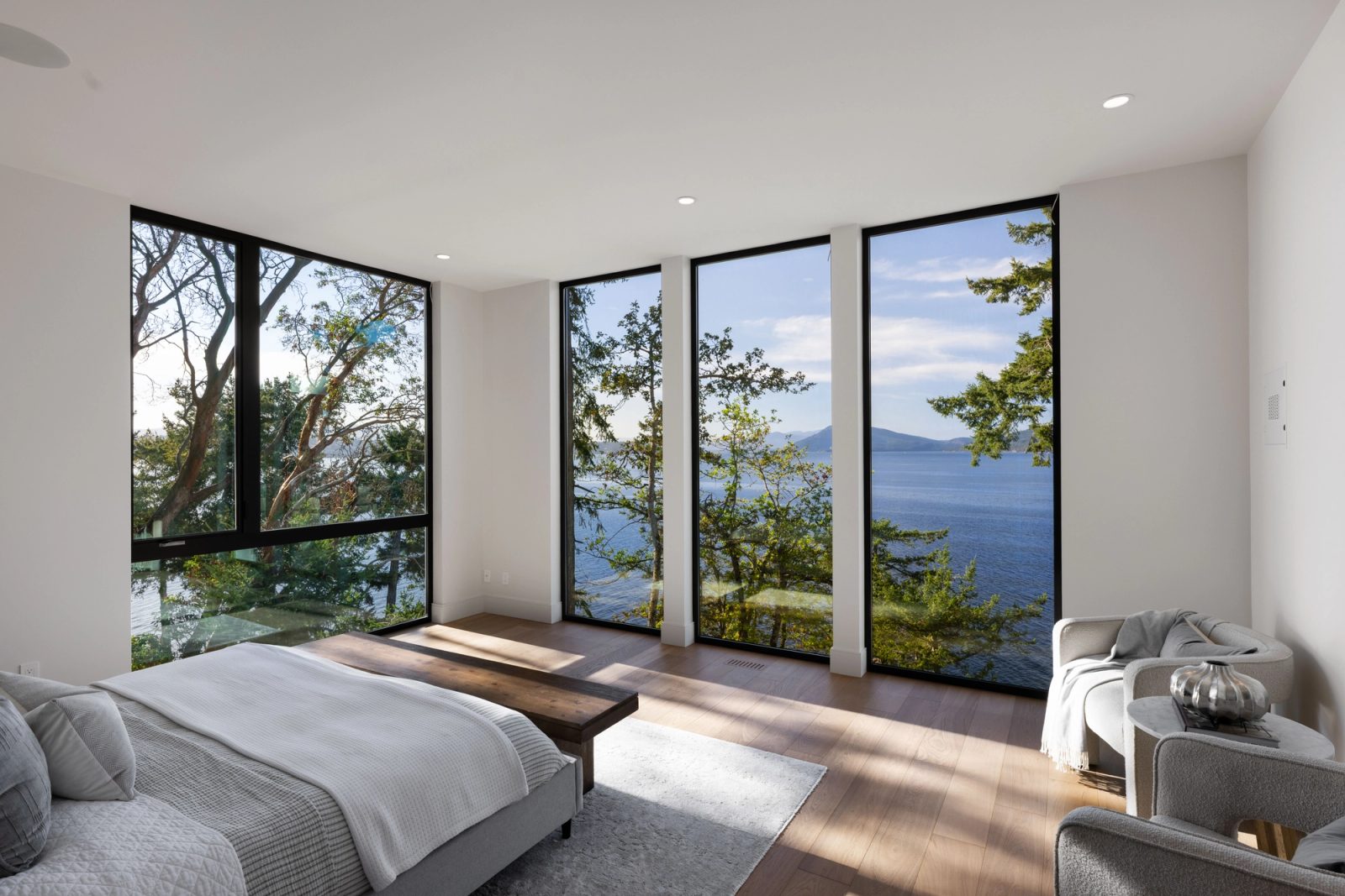 A bedroom with glazing on two sides, a low bed and bench facing tall windows divided by narrow piers, arbutus and open water beyond.