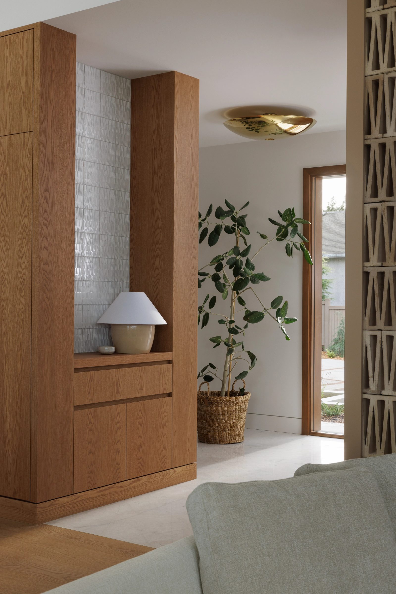 Left: an oak cabinet run beside a niche of white textured tile, a fig tree in a woven basket, and a screen of patterned block standing beyond it.