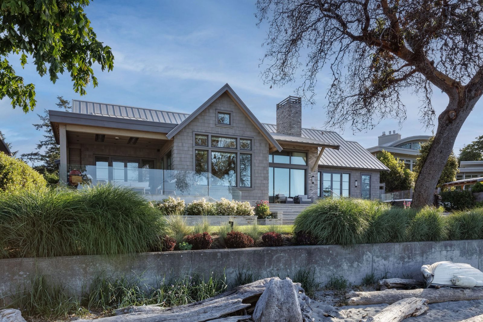 The cottage seen from the beach, framed by a Garry oak, with driftwood and a concrete seawall in the foreground