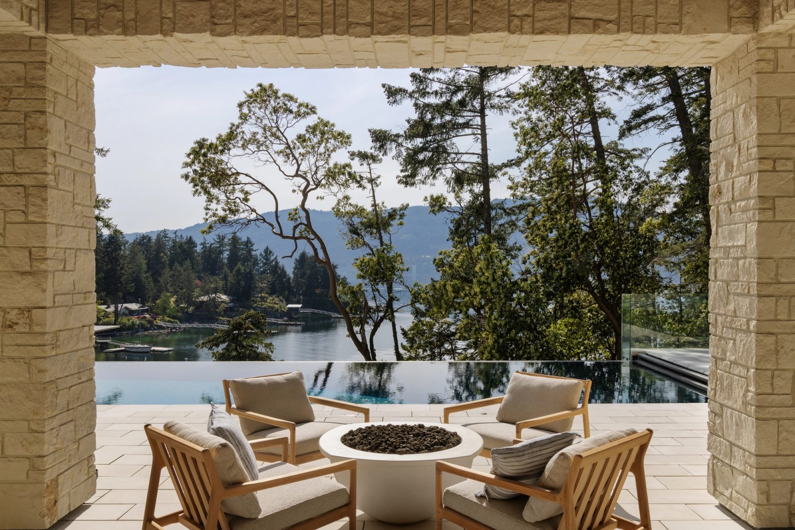 An infinity pool and spa at the edge of the drop, teak loungers alongside, the inlet and the far ridge past standing firs and arbutus.