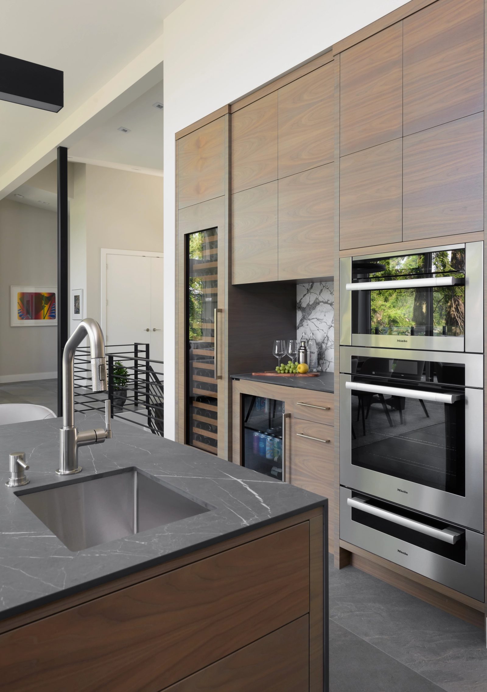 Kitchen island sink in dark veined stone with walnut cabinetry and stainless ovens beyond