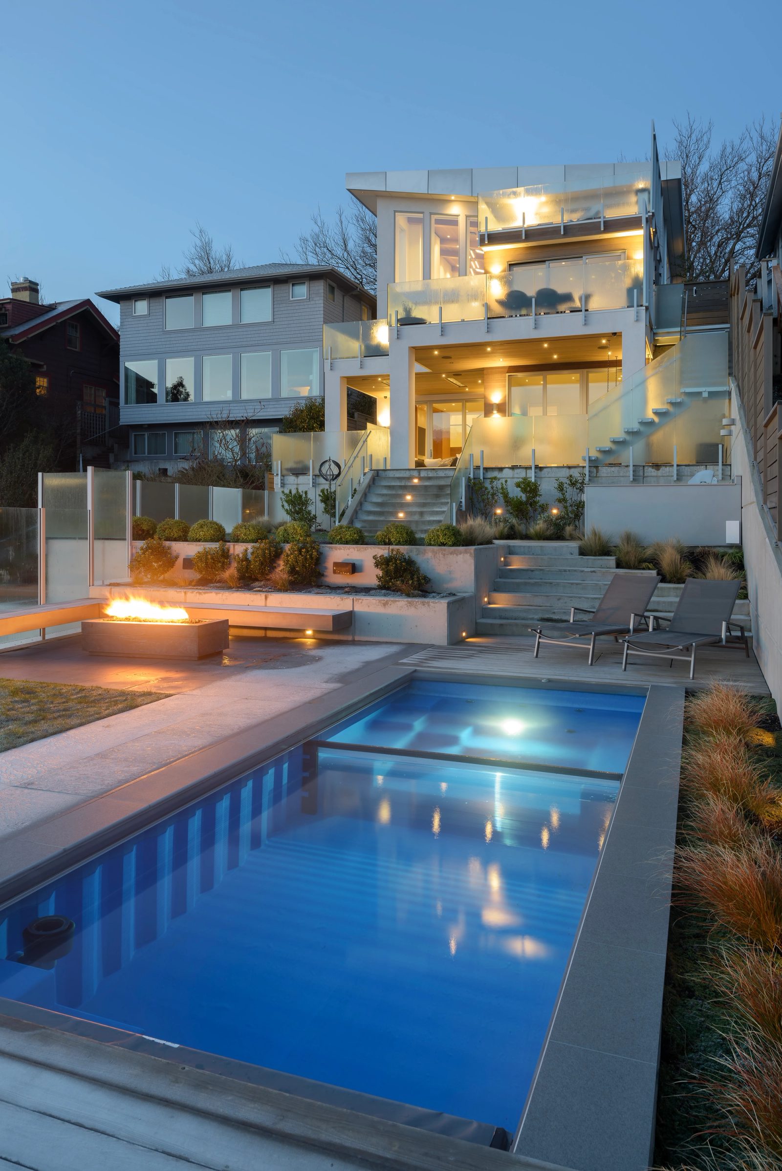Lap pool lit blue at dusk below a terraced garden, the house rising in lit tiers behind it.