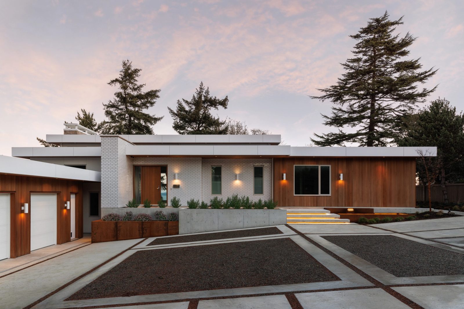 A long low house at dusk behind a forecourt laid in concrete panels and exposed aggregate, white brick and vertical cedar under flat roof planes, firs standing behind.