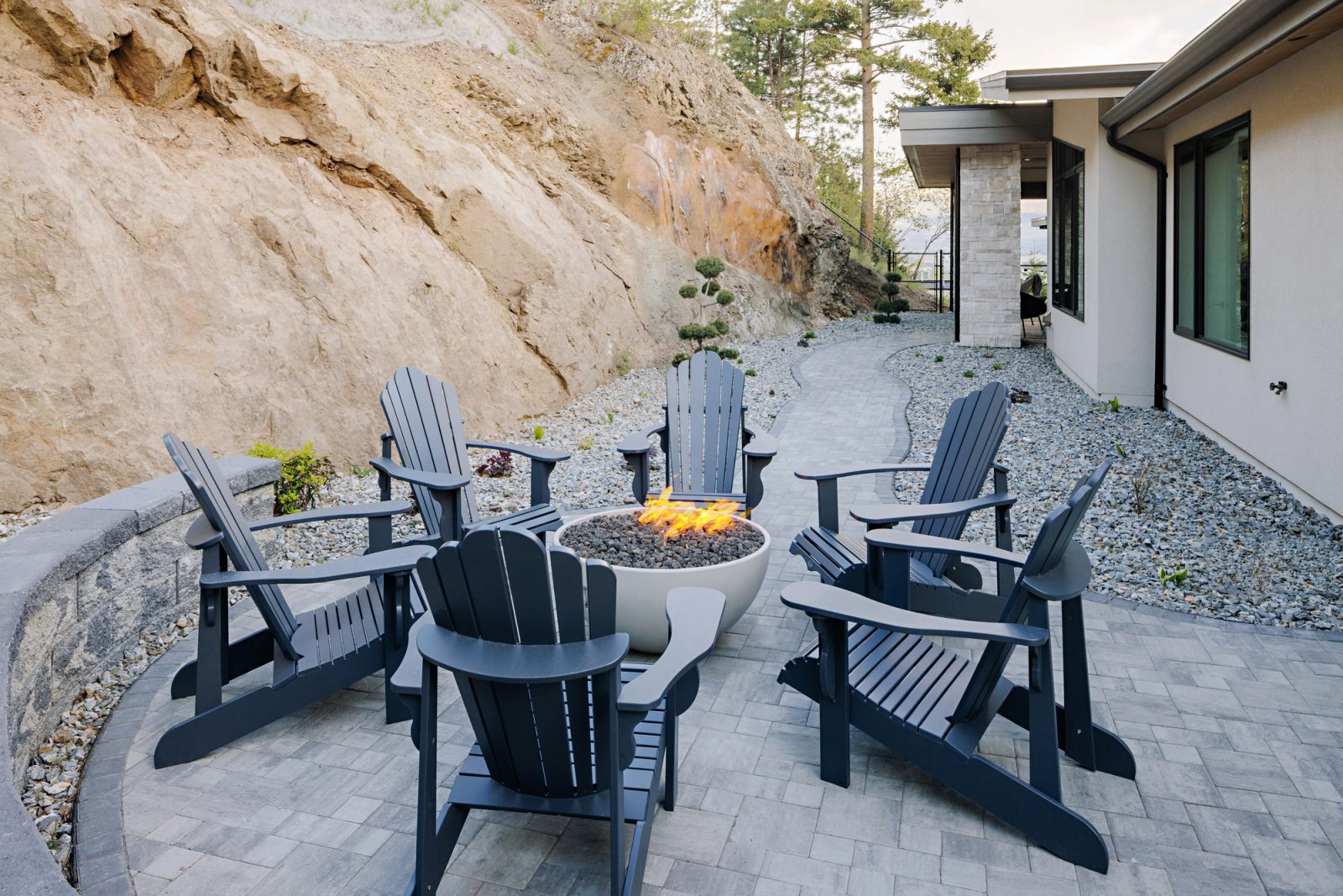 Six dark Adirondack chairs drawn around a low bowl fire pit on grey pavers, the cut rock face filling the frame behind them at last light.